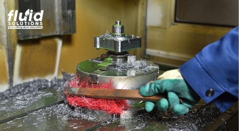 Worker cleaning metal shavings from machine component with brush.