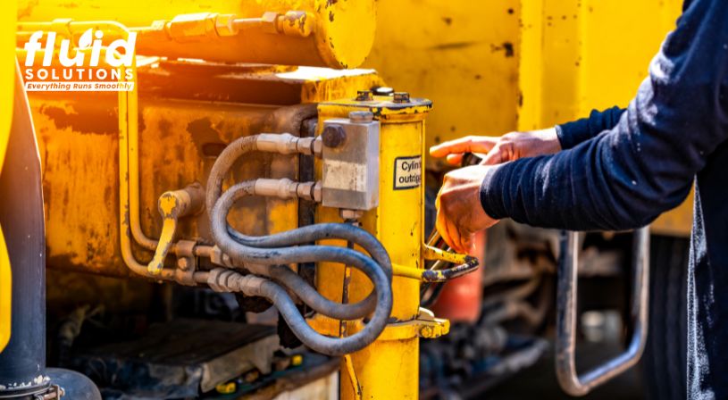 Worker operating a hydraulic system with multiple hoses on a yellow industrial machine.