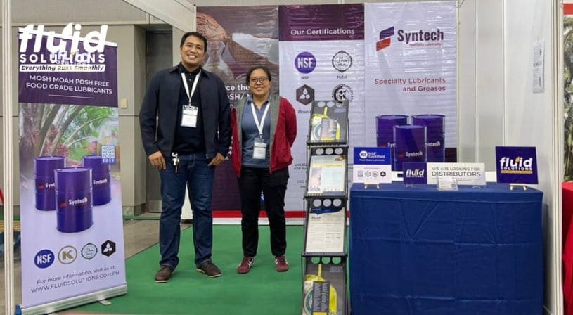 Fluid Solutions representatives posing together at a booth with Syntech lubricant products on display.