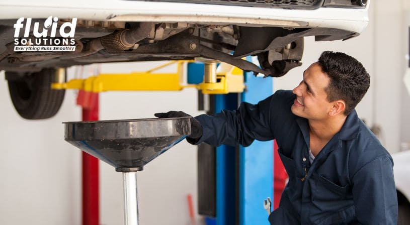 Mechanic smiling while performing an oil change on a car, with the Fluid Solutions logo visible.